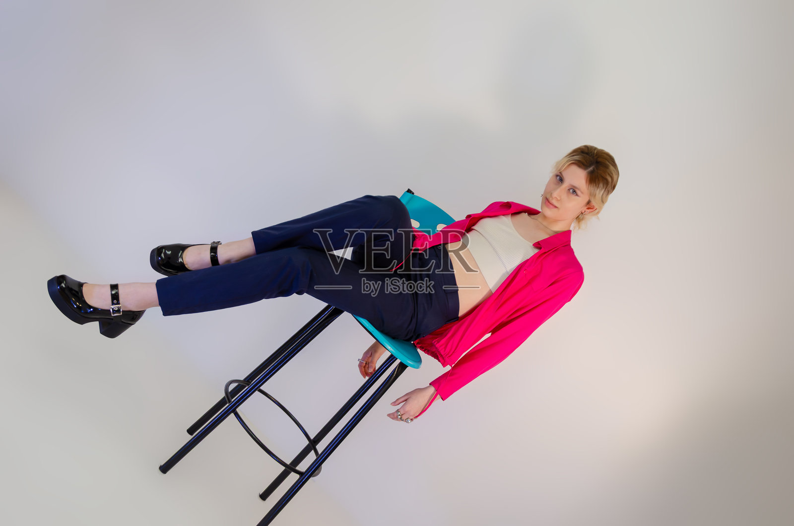 穿着粉色上衣和短款上衣的金发年轻女性自信地坐在椅子上，背景为白色。照片摄影图片