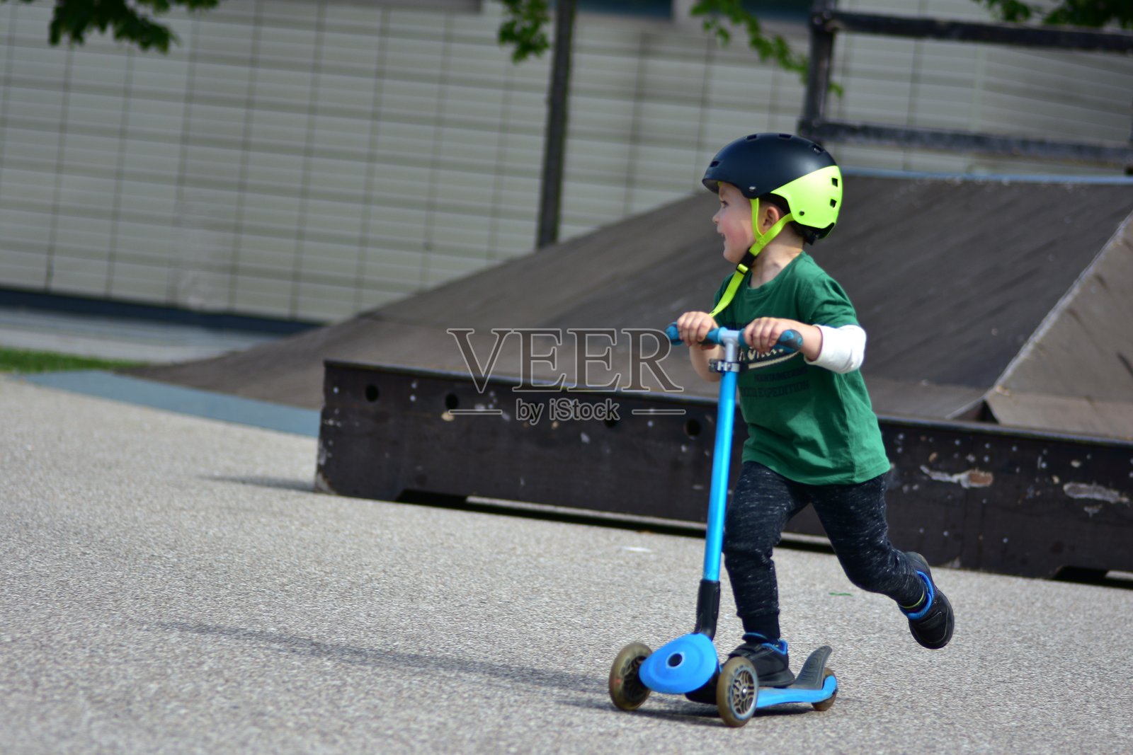活力四射的男孩穿着绿色衬衫和头盔，在滑板公园骑着蓝色三轮滑板车，面带快乐的表情，快速推动，享受速度、平衡和阳光明媚的户外游戏。照片摄影图片