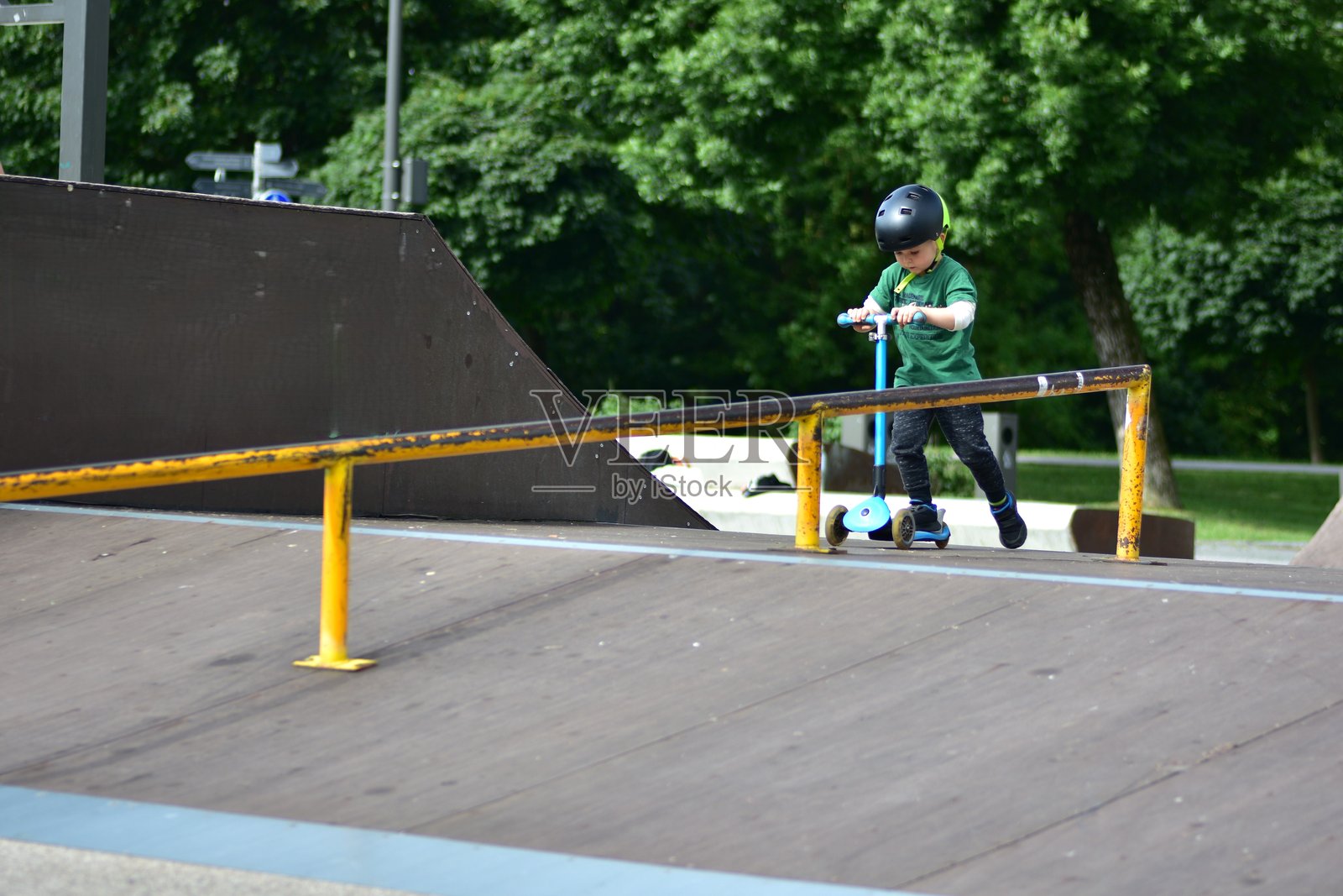 年轻男孩戴着黑色头盔，在滑板公园骑着蓝色三轮滑板车，专注地沿着滑板坡道前行。照片摄影图片