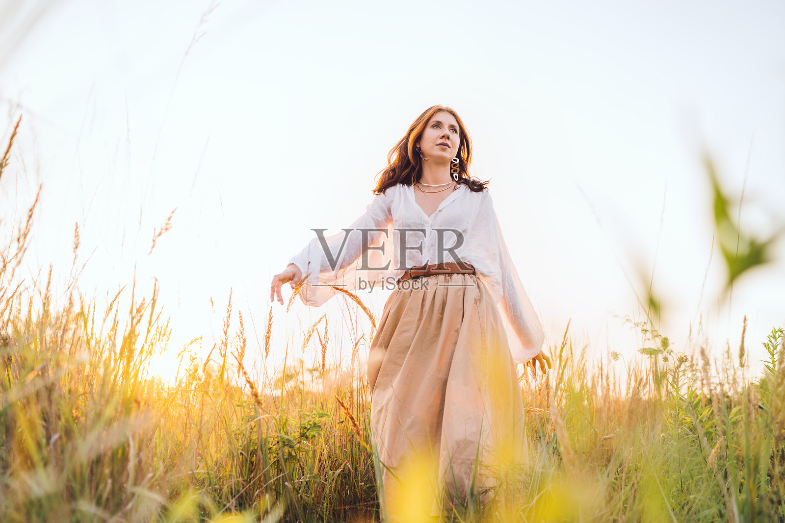 年轻女性穿着白色衬衫和米色裙子，在金色草地上自由地漫步，享受温暖的日落阳光，感受宁静的自然氛围，表达自由、宁静、正念与自然的和谐。照片摄影图片