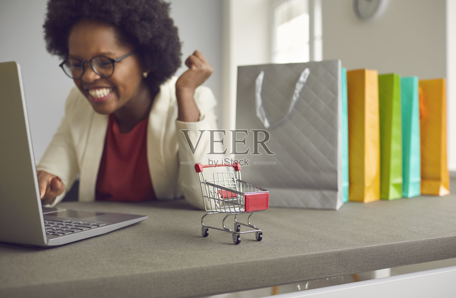 特写镜头：一辆小购物车放在桌子上，背后是一位肤色较深的女性正在网上购物。照片摄影图片