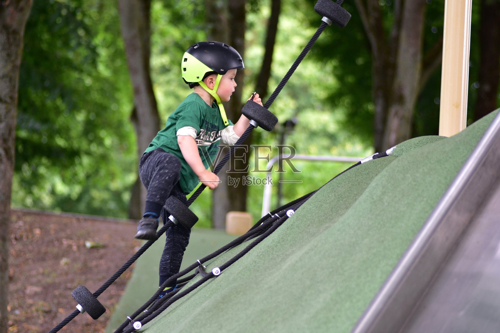年轻男孩戴着头盔在游乐场的攀爬山上爬绳梯，展现出在绿色户外设施上积极玩耍时的决心和专注。照片摄影图片