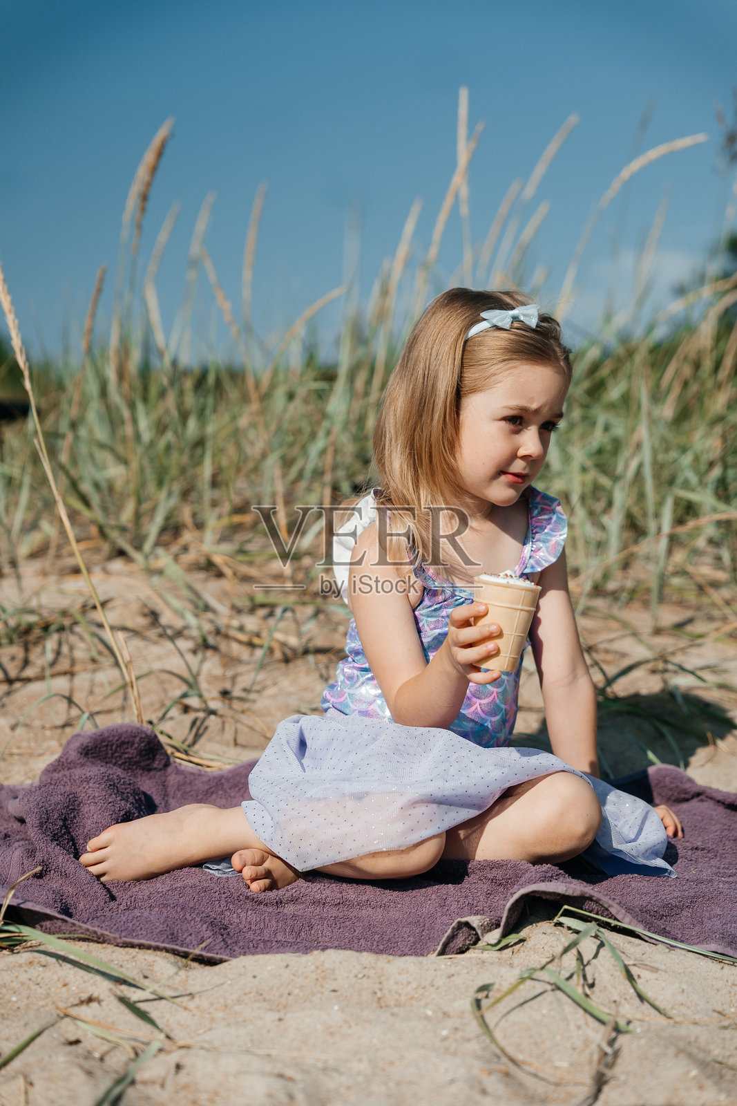 孩子在阳光明媚的日子里，享受着冰淇淋，坐在沙滩上，周围是高高的草丛。照片摄影图片