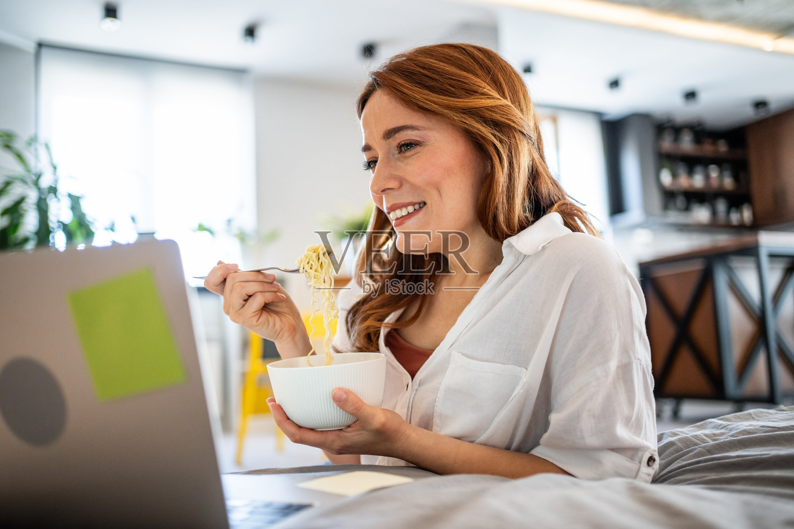微笑的女人在床上吃面条并观看笔记本电脑上的电影照片摄影图片