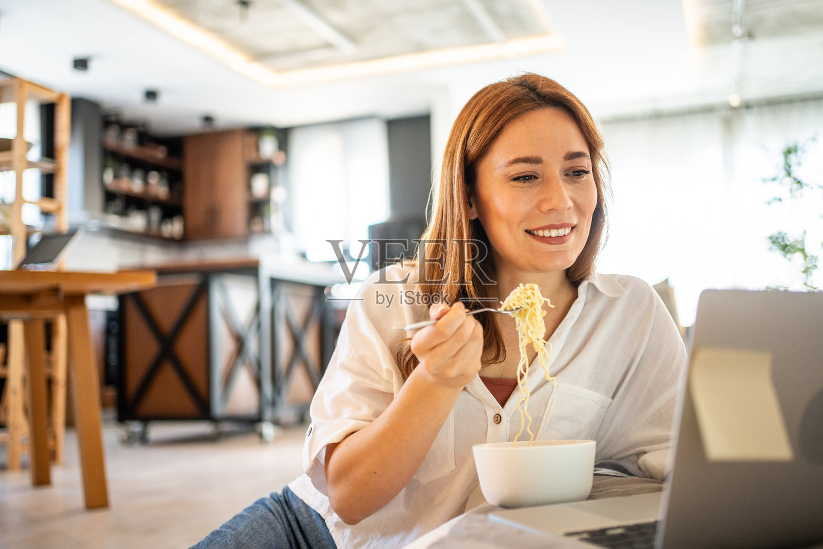 年轻女性在家工作，一边吃面条一边看笔记本电脑照片摄影图片