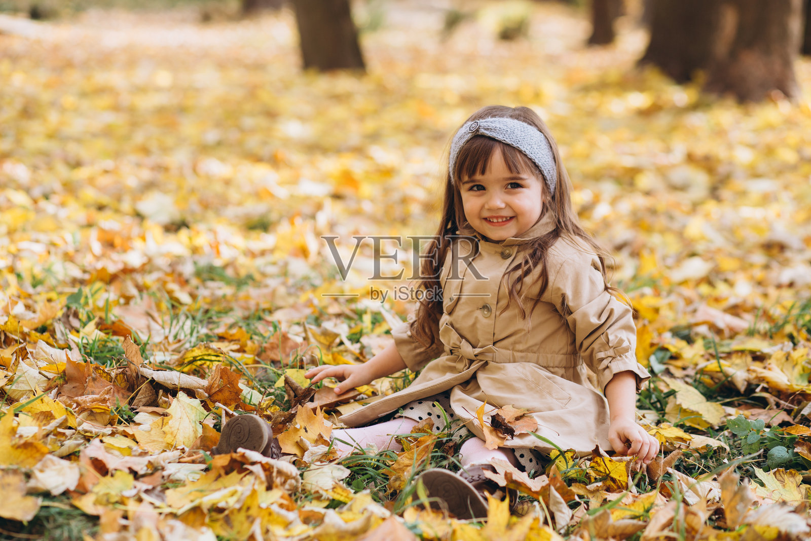 快乐而美丽的小女孩穿着米色外套，坐在秋天公园的黄色落叶中。照片摄影图片
