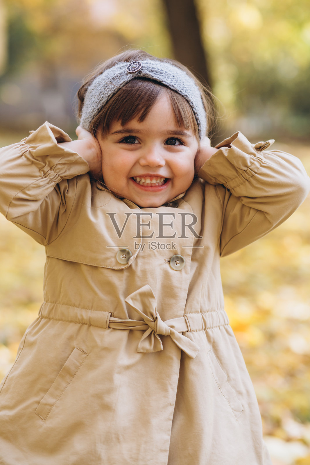 快乐而美丽的女孩穿着米色外套，在秋天的公园里表现出情感，用手指捂住耳朵。照片摄影图片