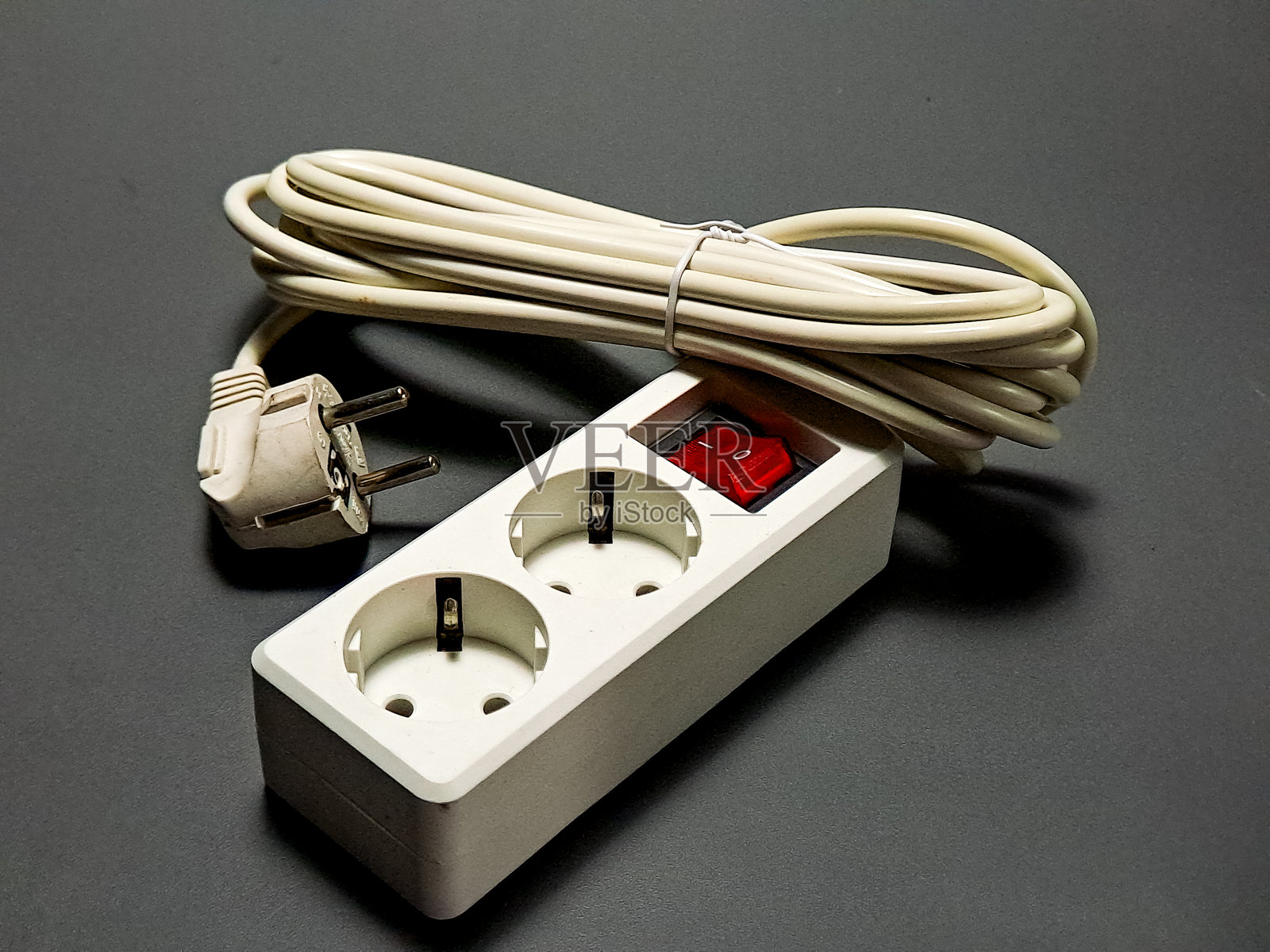 一个带有两个插座和卷曲电线的白色插线板放在灰色表面上，红色开关亮起，表示电流流动，这是一个常见的电力分配设备。照片摄影图片