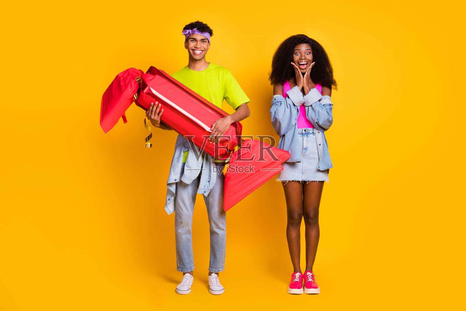 一对时尚快乐的情侣手持大型假糖果，站在明亮的黄色背景前，展现全身形象。照片摄影图片