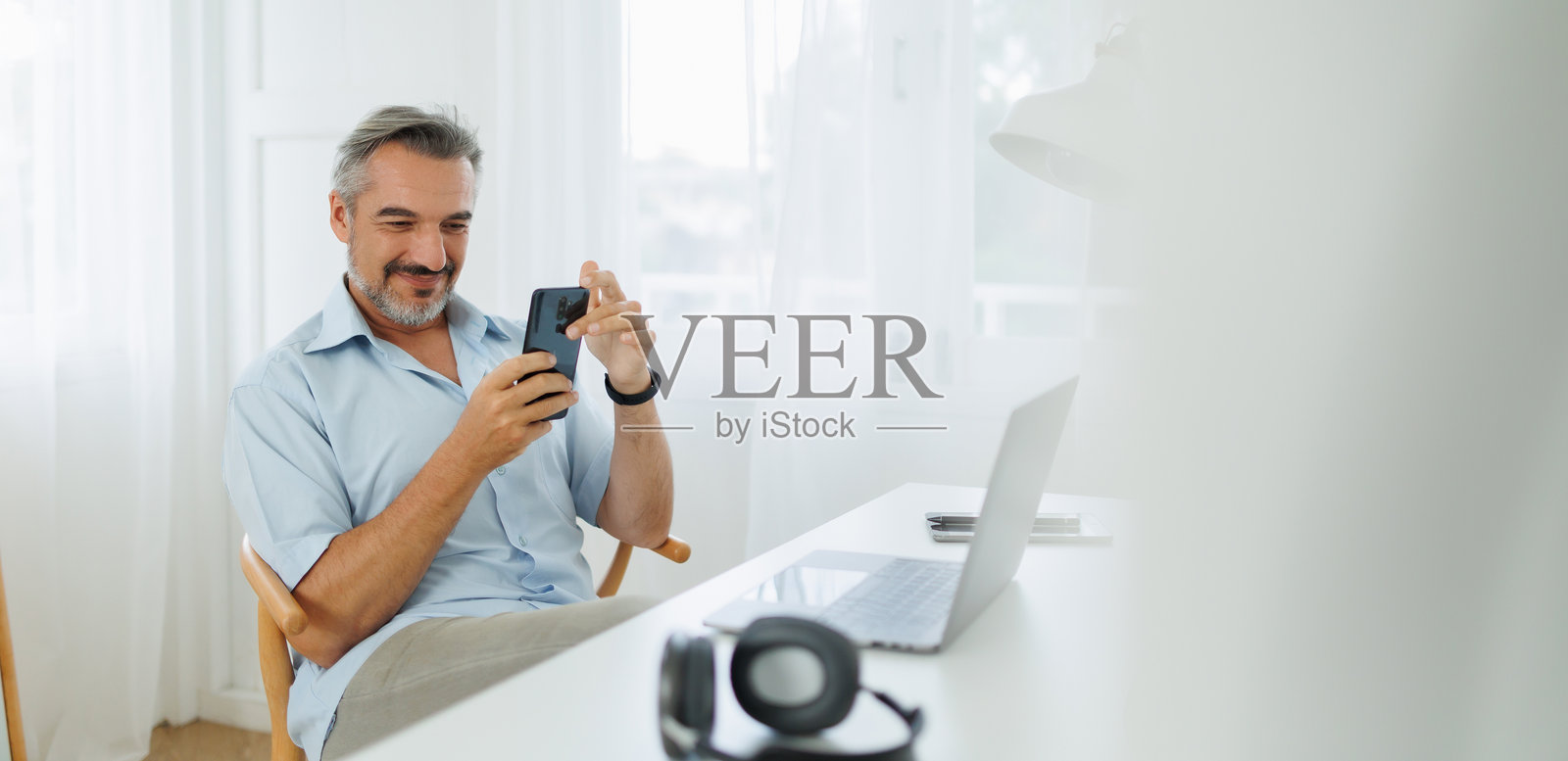 中年男子在整洁的家庭办公室里微笑着使用智能手机和笔记本电脑。照片摄影图片