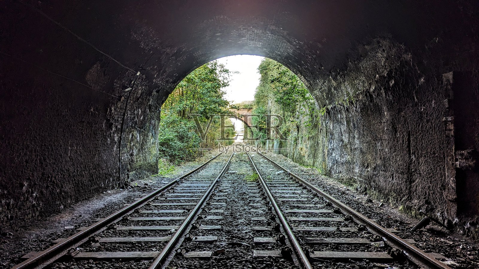 隧道尽头的光线，透过石拱门可见树木和铁路桥，废弃的双轨长铁轨和拱门墙壁上投射着黑暗的阴影。照片摄影图片