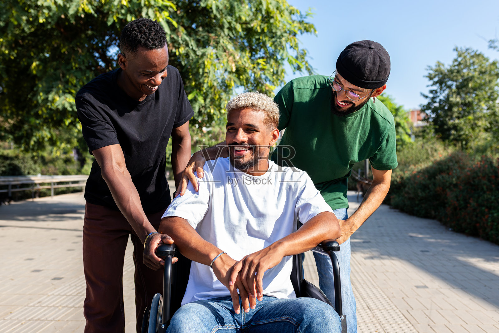两个朋友在公园里推着他们的残疾朋友坐在轮椅上。照片摄影图片