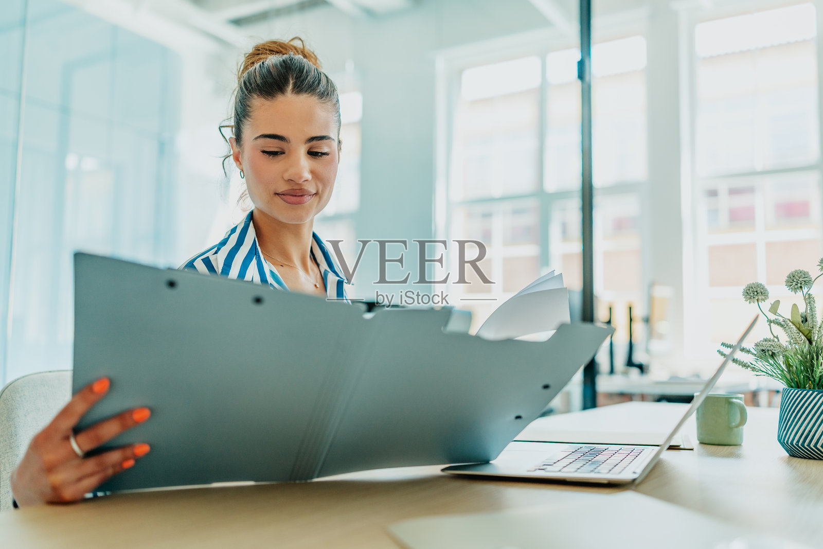 年轻女性在明亮的办公室里愉快地工作，边查看文件边对着笔记本电脑微笑。照片摄影图片