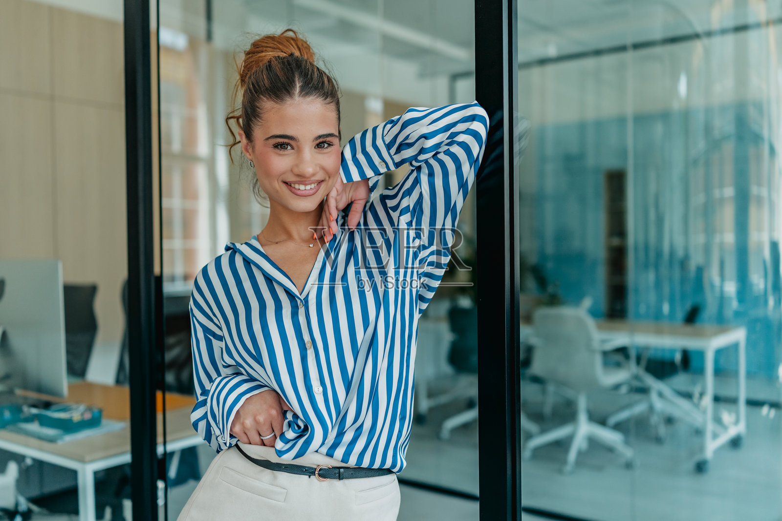 微笑的年轻女性穿着条纹衬衫，在明亮现代的室内工作空间中摆姿势，正值白天。照片摄影图片