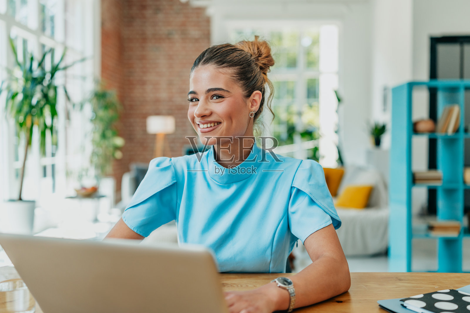 年轻女性在明亮的客厅中微笑着使用笔记本电脑，周围环绕着植物，营造出舒适的氛围。照片摄影图片