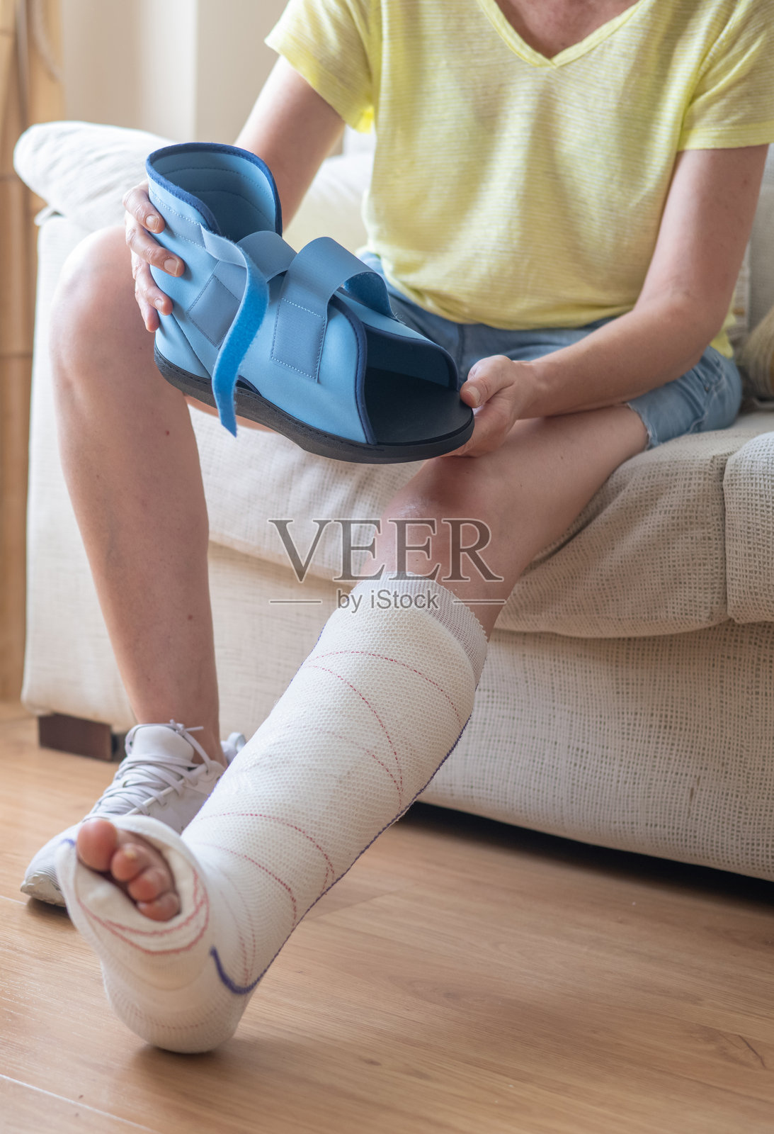 成熟的女性坐在沙发上，手里拿着矫形靴，刚刚接受了跖骨骨折手术。她穿着石膏，专注于在家中的康复。照片摄影图片
