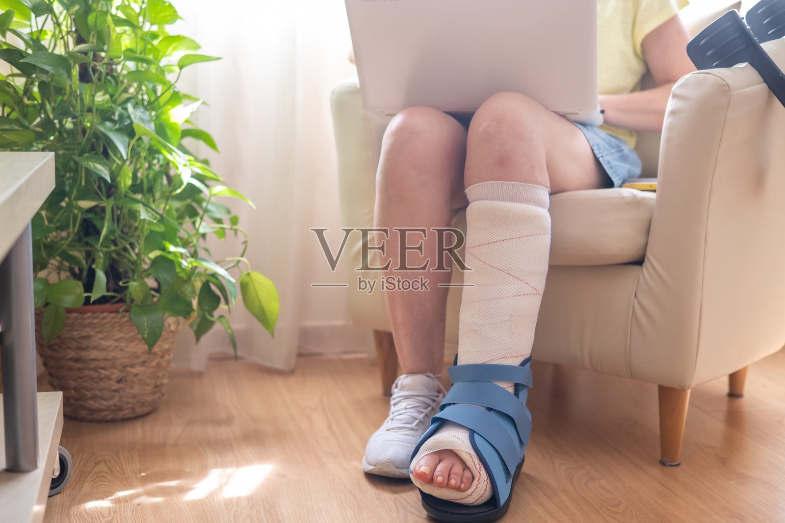 成熟的女性脚踝骨折，穿着医疗靴，舒适地坐在扶手椅上使用笔记本电脑，专注于康复并适应家庭中的新挑战。照片摄影图片