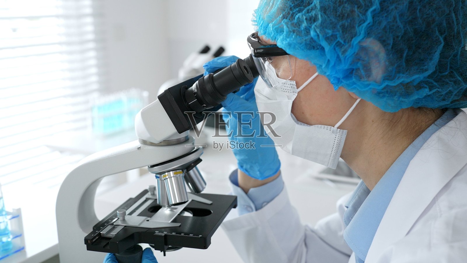 女性科学家戴着口罩和蓝色安全装备，正在使用显微镜进行研究，背景中有一位男性同事在工作。医学、医疗和科学概念照片摄影图片