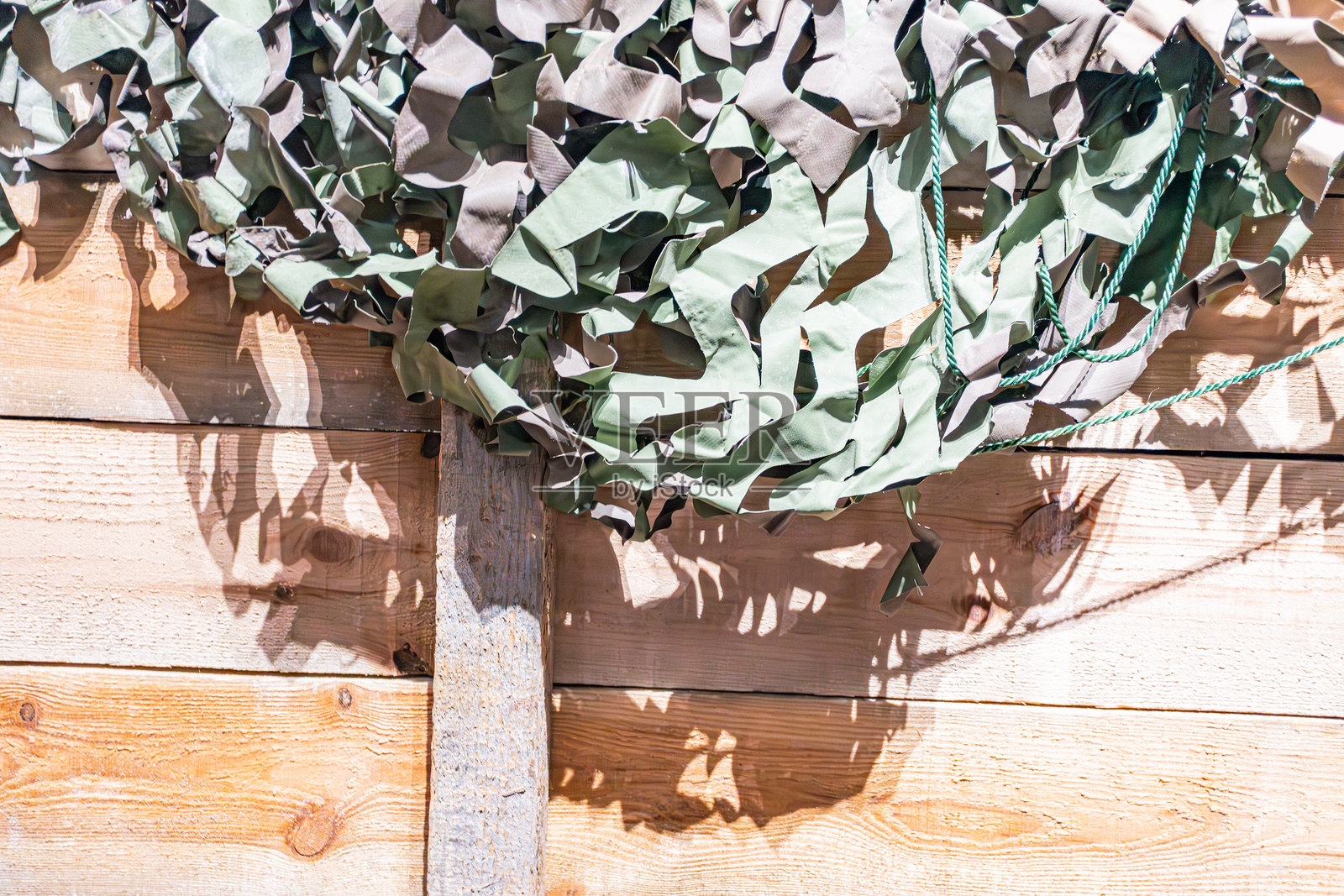 墙壁上挂着的光滑绿色阳光爆发图案的人工植物，放置在木板上，背景是乡村风格的木板墙，旁边有军事迷彩网，自然室内光线投射出阴影。照片摄影图片