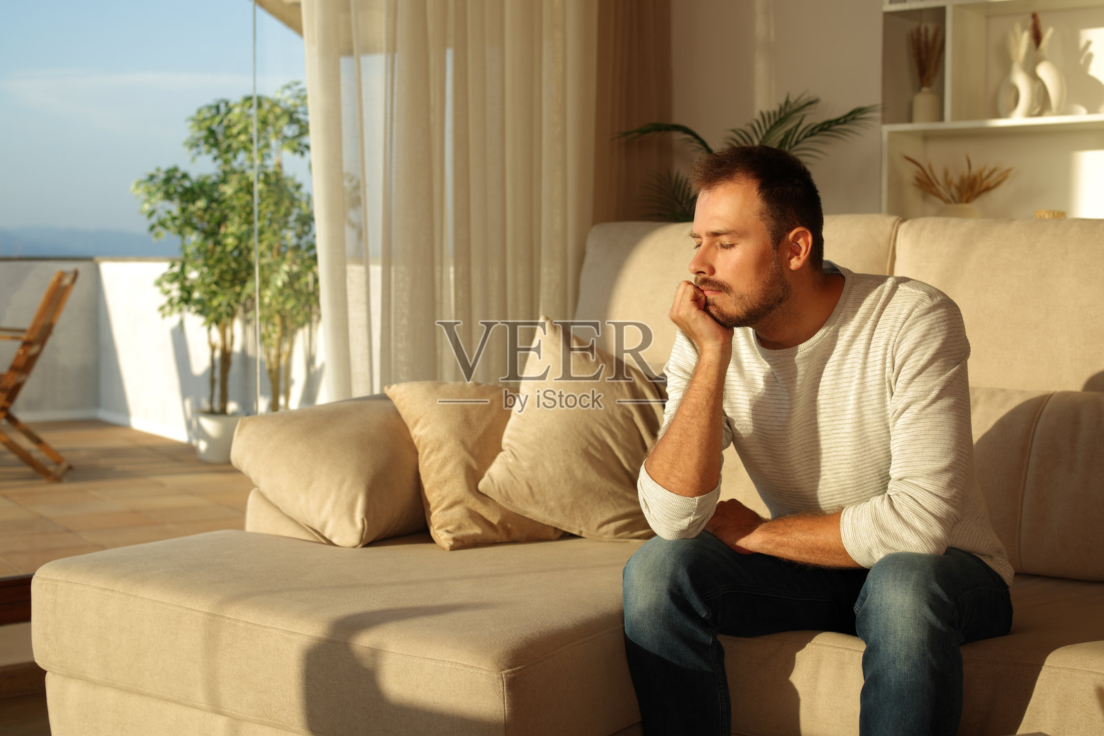 男人在家中日落时坐在沙发上放松，闭着眼睛。照片摄影图片