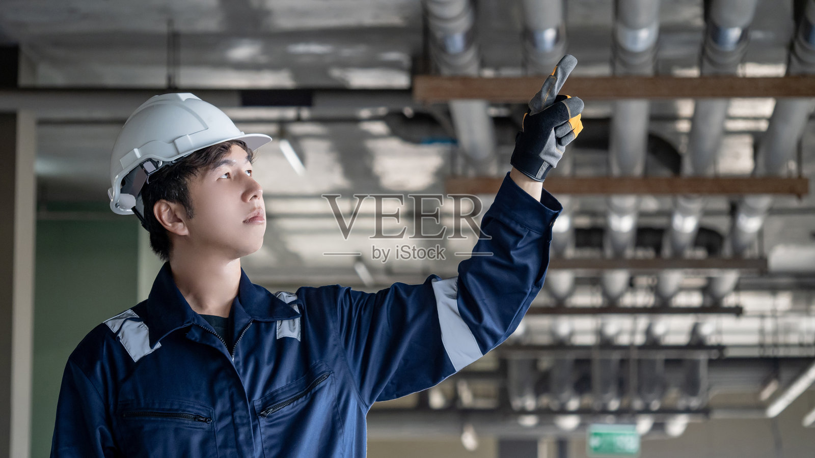 自信的亚洲男性建筑工人穿着防护服和安全头盔，指着建筑中的管道系统照片摄影图片