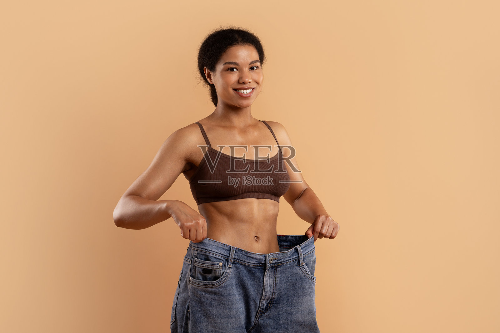 一位黑人女性穿着内衣，手持超大号牛仔裤以展示减肥进展。照片摄影图片