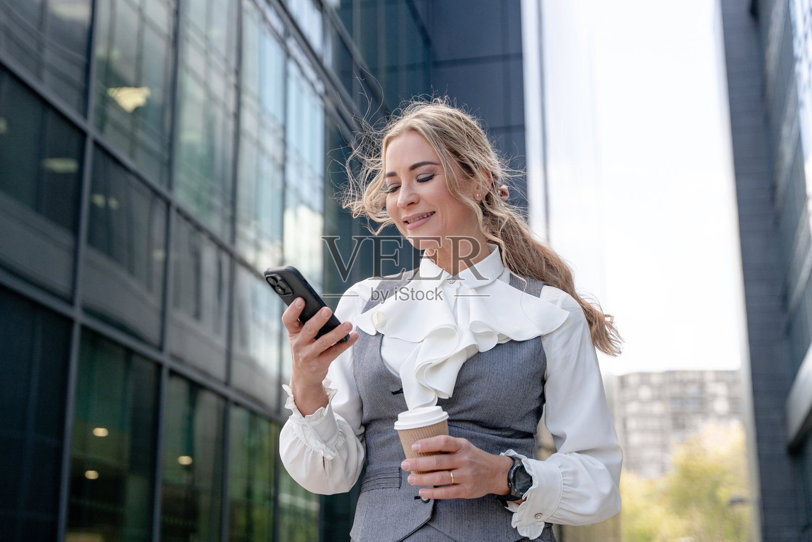 女商人在阳光明媚的日子里，走在城市街道上，一边喝咖啡一边发短信，身旁是现代玻璃建筑。照片摄影图片
