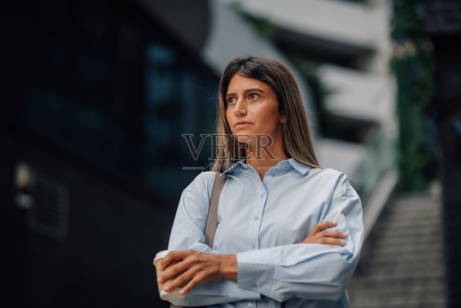 一位商务女性在现代办公大楼前双臂交叉，手中拿着外带咖啡，正在等待。照片摄影图片