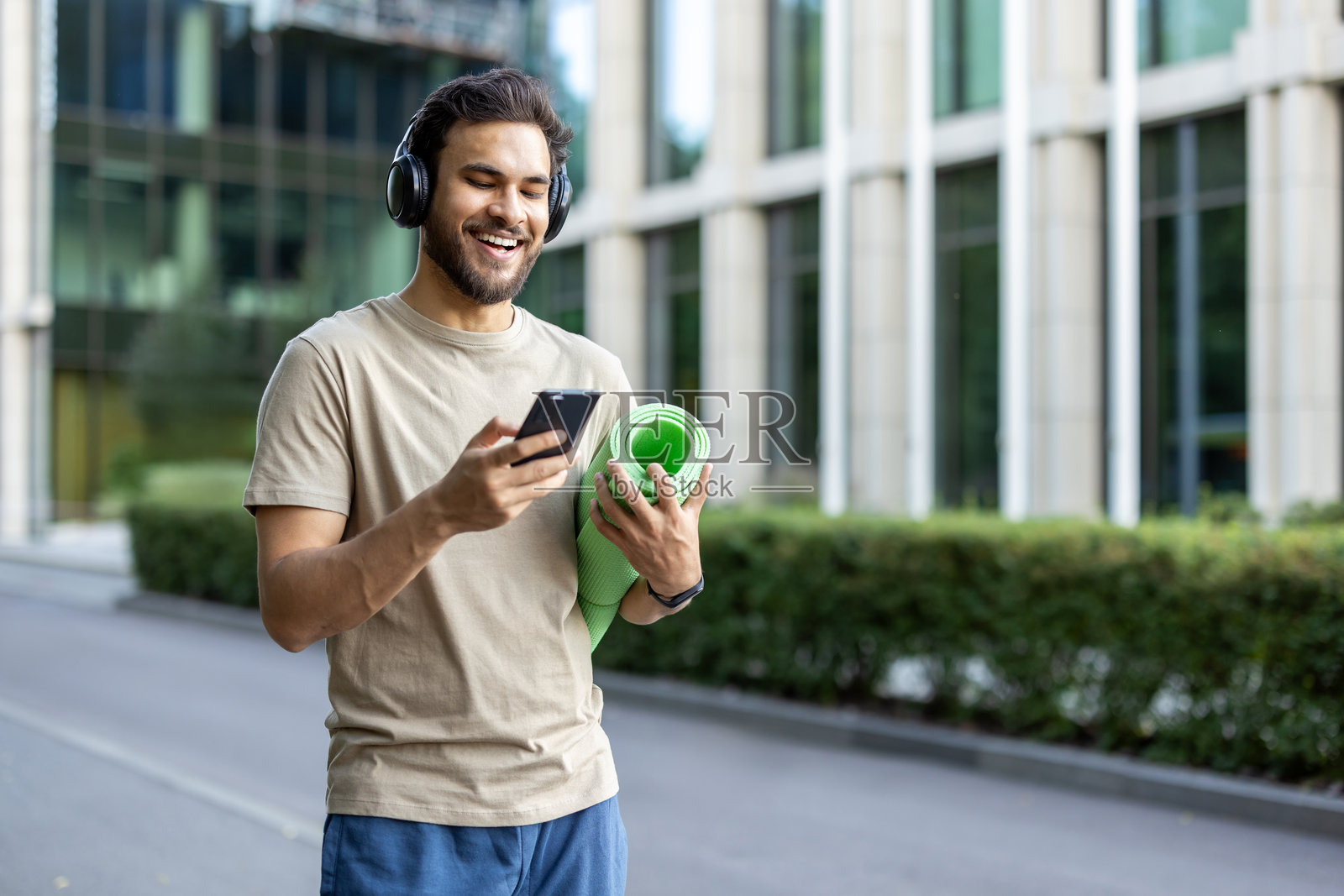 一位微笑的年轻印度男子在户外运动，戴着耳机，手里拿着手机和瑜伽垫。照片摄影图片