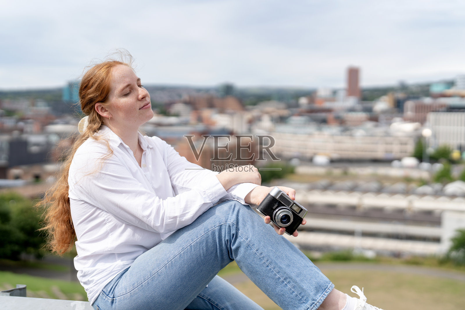 年轻女性坐在窗台上，手持相机，享受阴天城市天际线的美景。照片摄影图片