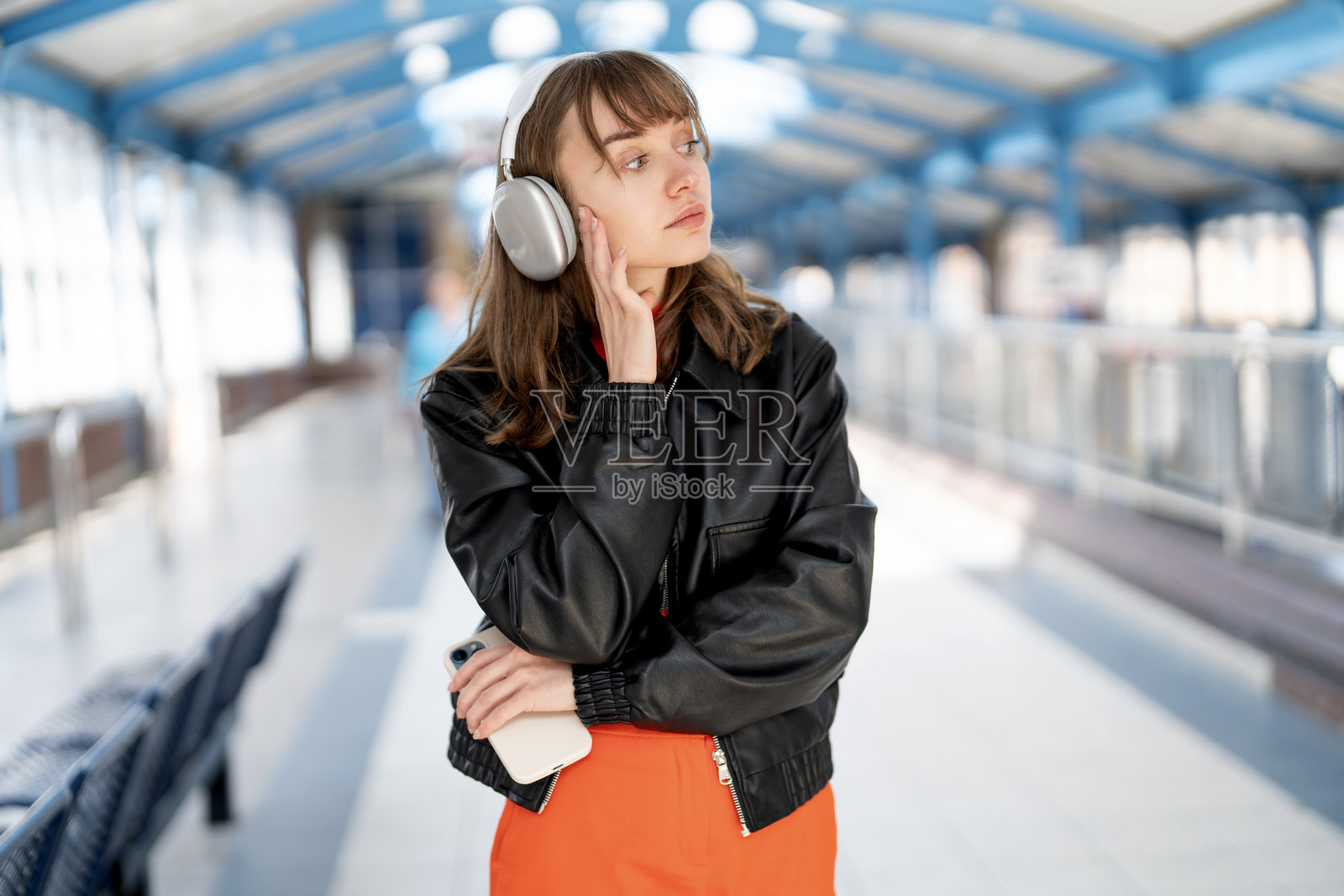 年轻女性戴着耳机，白天在城市交通枢纽思考，同时查看手机。照片摄影图片