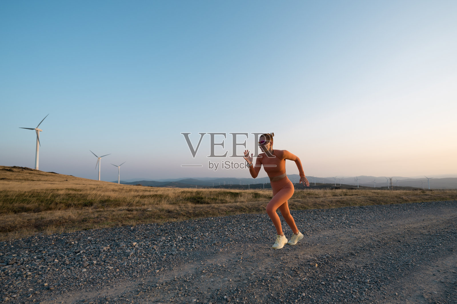 年轻女性在日落时分沿着风力发电机旁的碎石路跑步照片摄影图片
