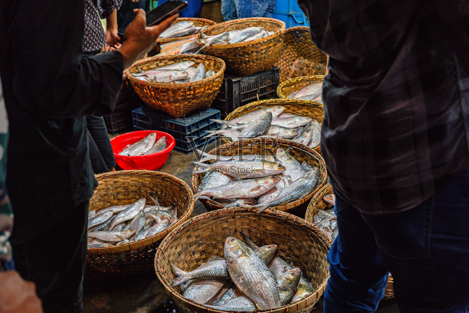 希尔萨鱼，南亚的一种美味，在孟加拉国达卡的当地市场出售。这些鱼被放在冰上，装在传统的柳条篮子和蓝色塑料箱中，准备出售给顾客。市场热闹非凡，充满了活力。照片摄影图片