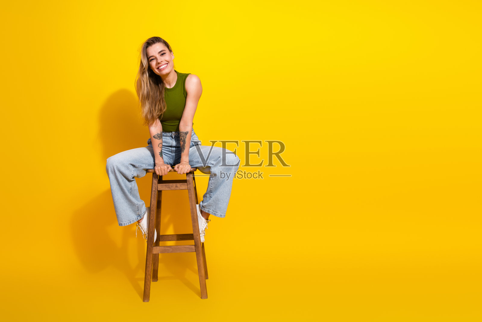 微笑的年轻女性穿着休闲时尚的服装，坐在凳子上，背景是明亮的黄色。照片摄影图片