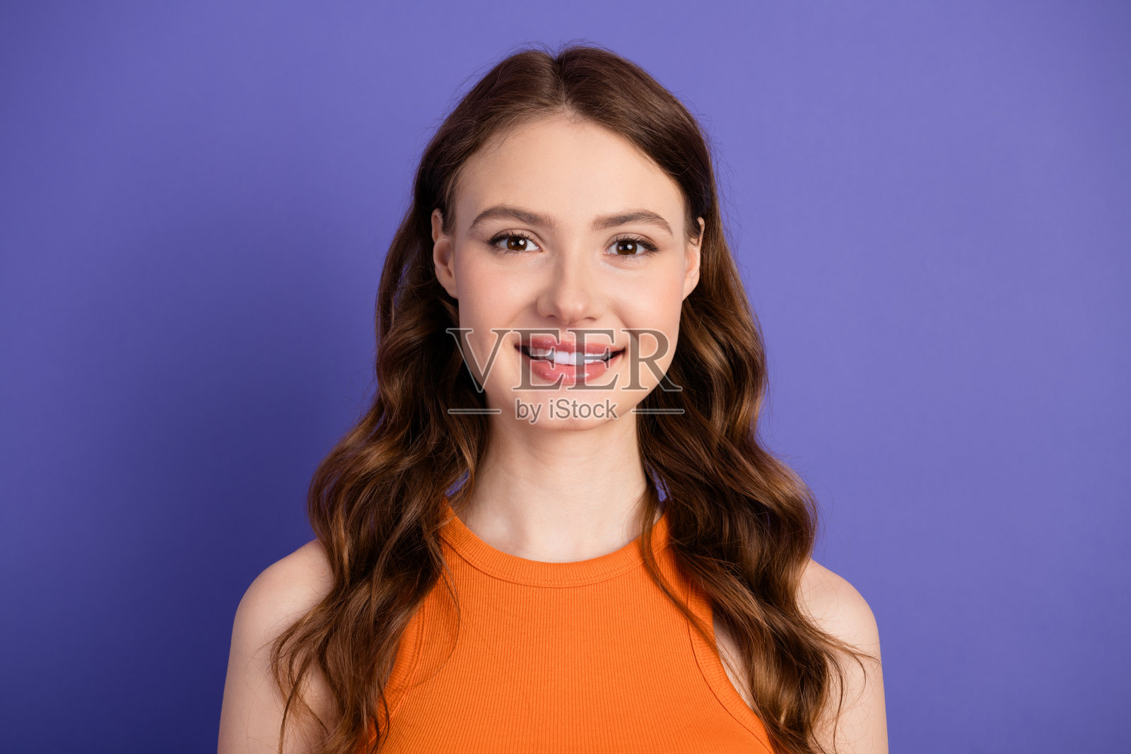 可爱时尚的年轻女性，拥有迷人的长棕色头发，穿着橙色上衣，站在鲜艳的紫色背景前。照片摄影图片