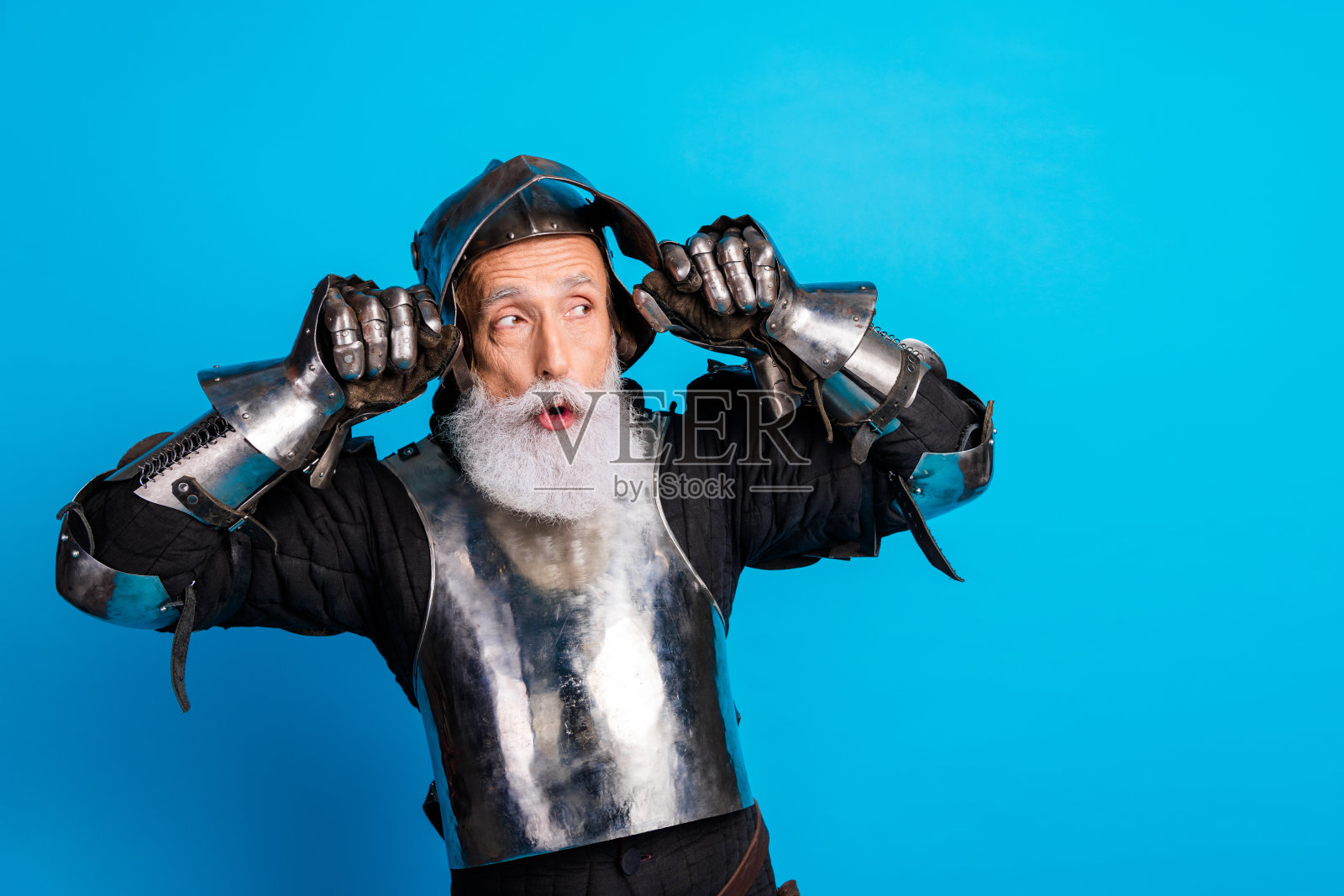身穿闪亮盔甲的老年男子在生动的蓝色背景前幽默地摆姿势，体现出高贵与滑稽的结合。照片摄影图片