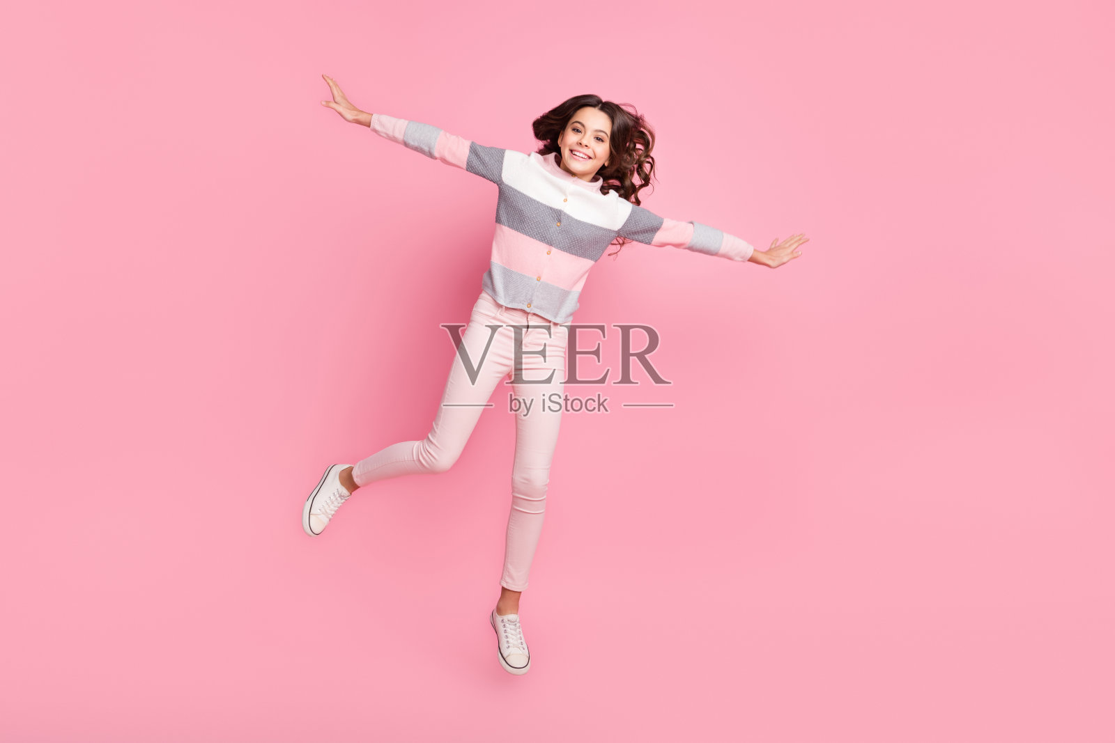 全身照，吸引人的充满活力的年轻女孩在粉色背景上跳起，双手做出翅膀的姿势。照片摄影图片