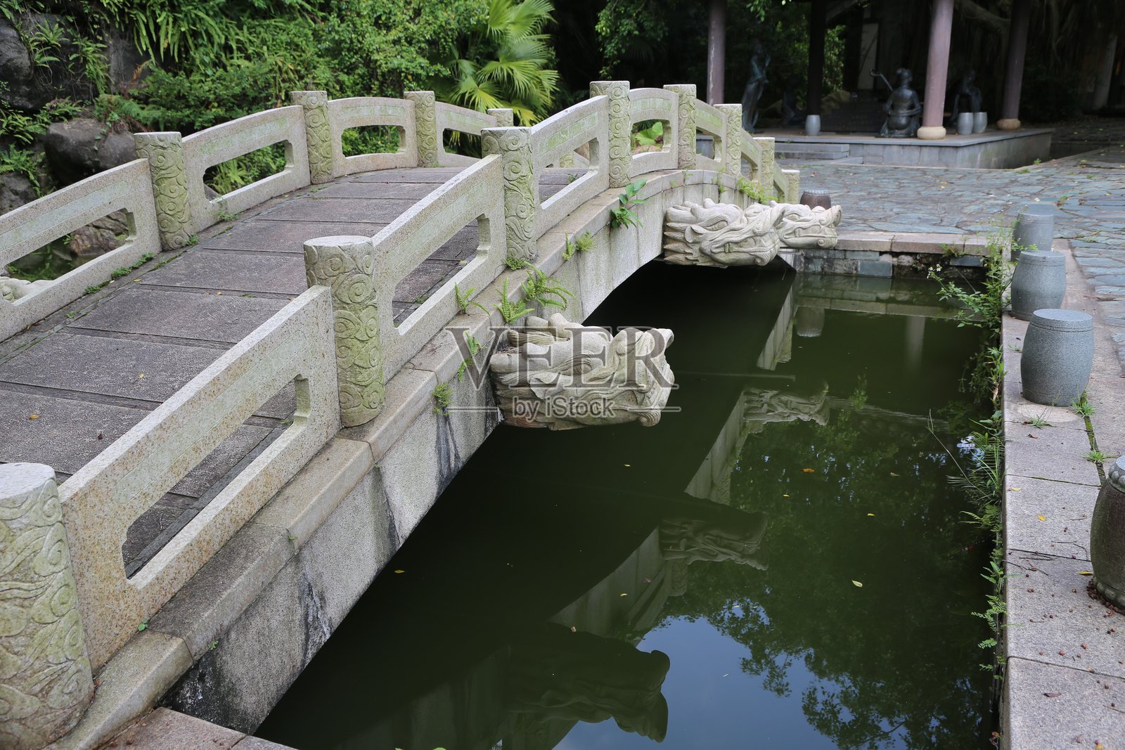 一座带有龙头栏杆的石桥通向前方的小池塘，池塘四周环绕着绿色植物和树木。照片摄影图片