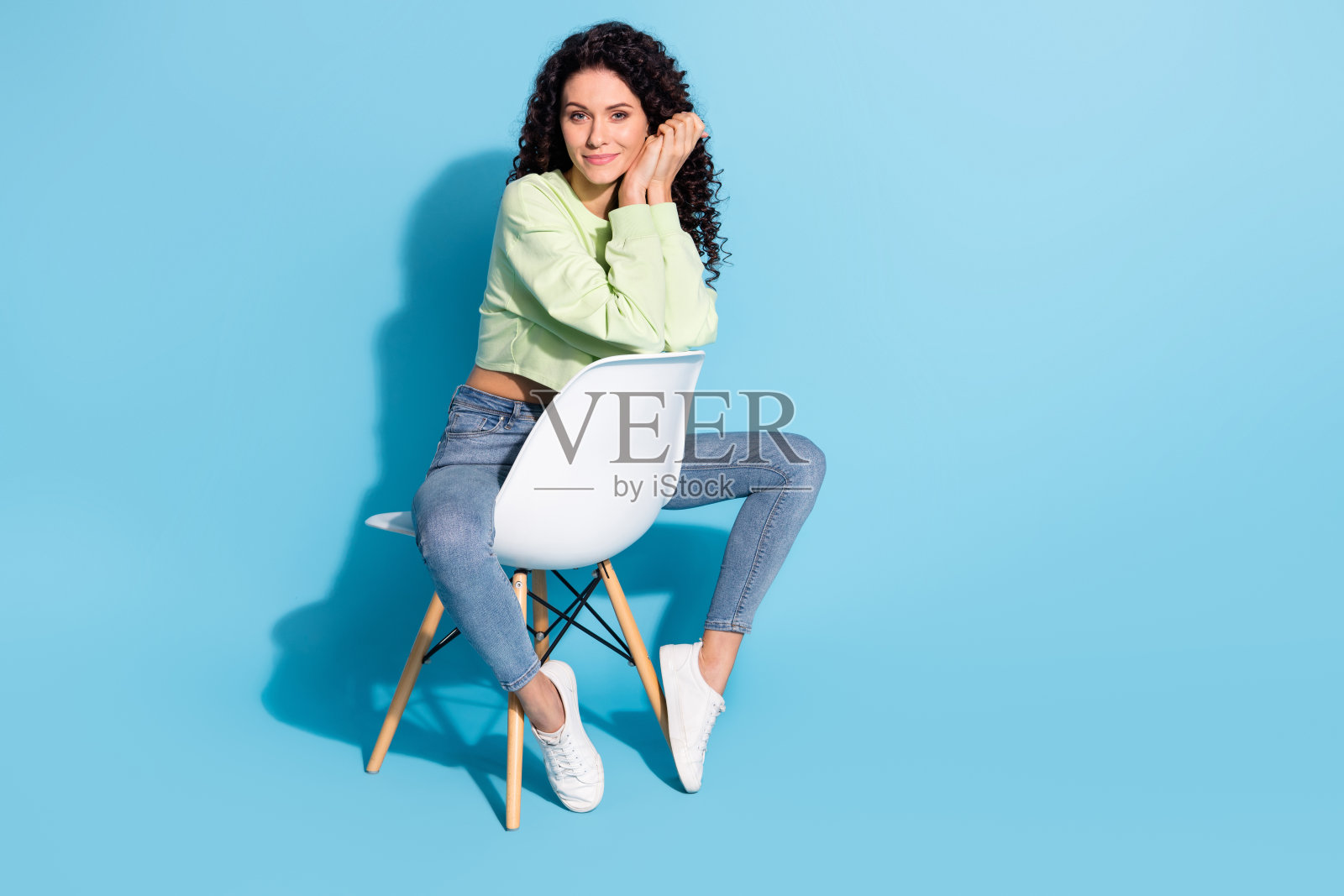 可爱满意的女士全身肖像，双臂交叉，手掌托腮，面向镜头，背景为蓝色。照片摄影图片