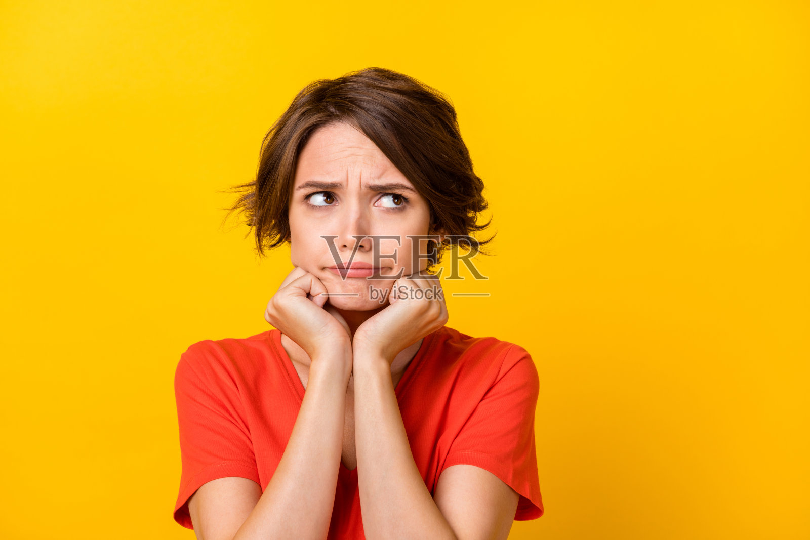 一位可爱的棕发女士，双手托着脸颊，穿着红色T恤，背景为鲜艳的黄色。照片摄影图片