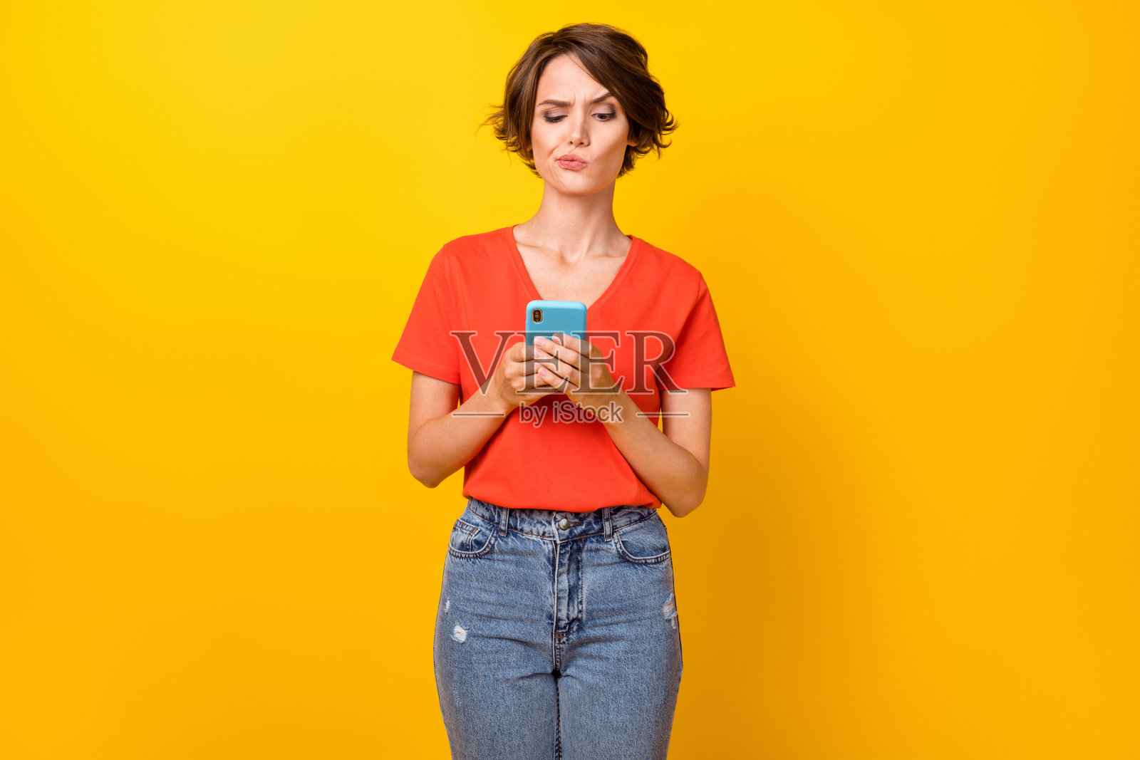 穿着红色T恤的漂亮棕发女士悲伤地看着电话，背景是鲜艳的黄色。照片摄影图片