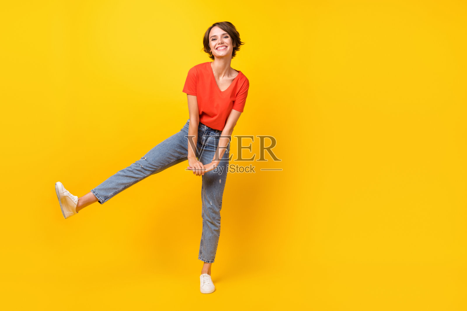 全身照，酷炫的棕发乐观女孩穿着红色T恤、牛仔裤和运动鞋，背景为黄色。照片摄影图片