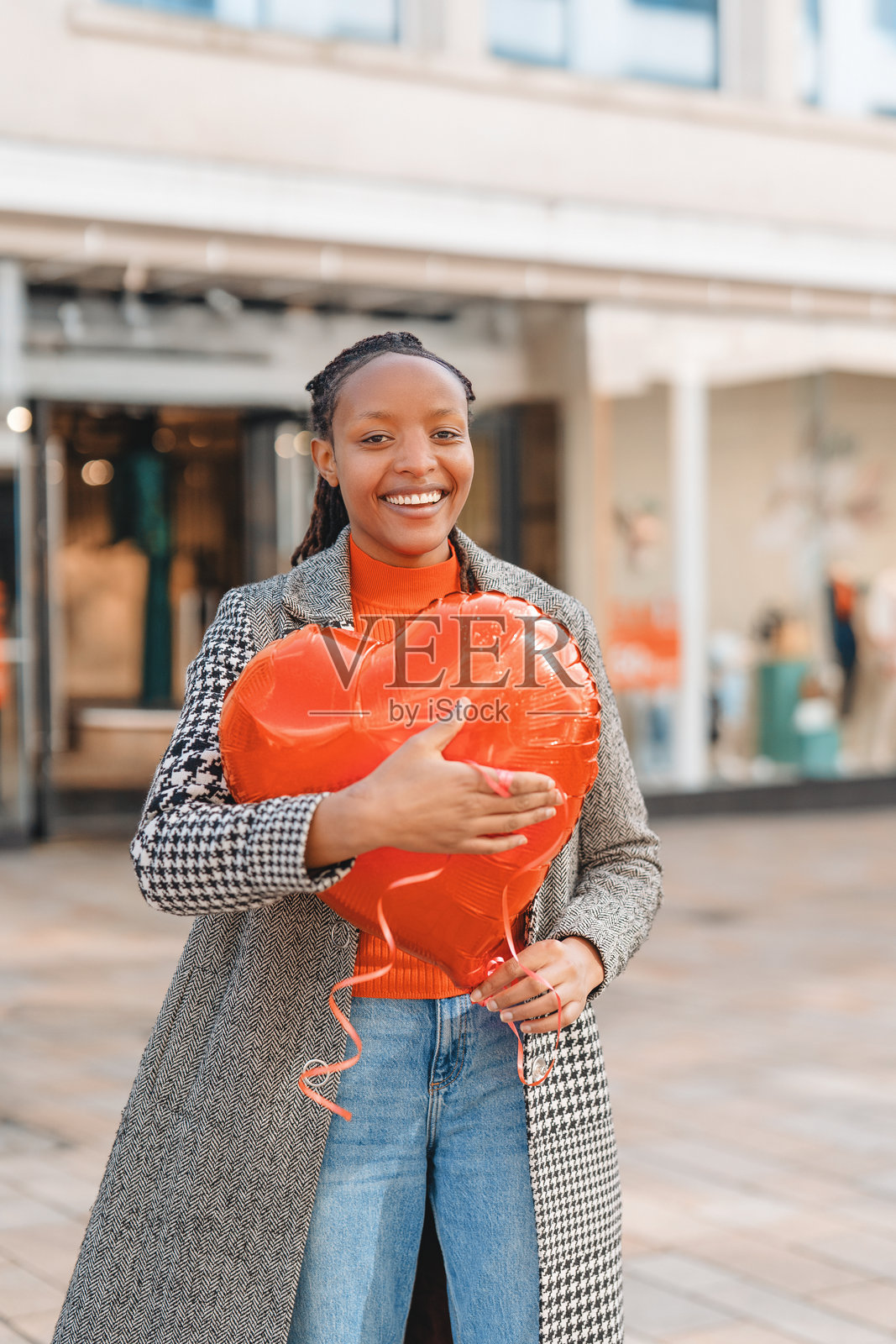 年轻女子在户外购物区微笑，手中拿着一个大型红色心形气球，阳光明媚。照片摄影图片