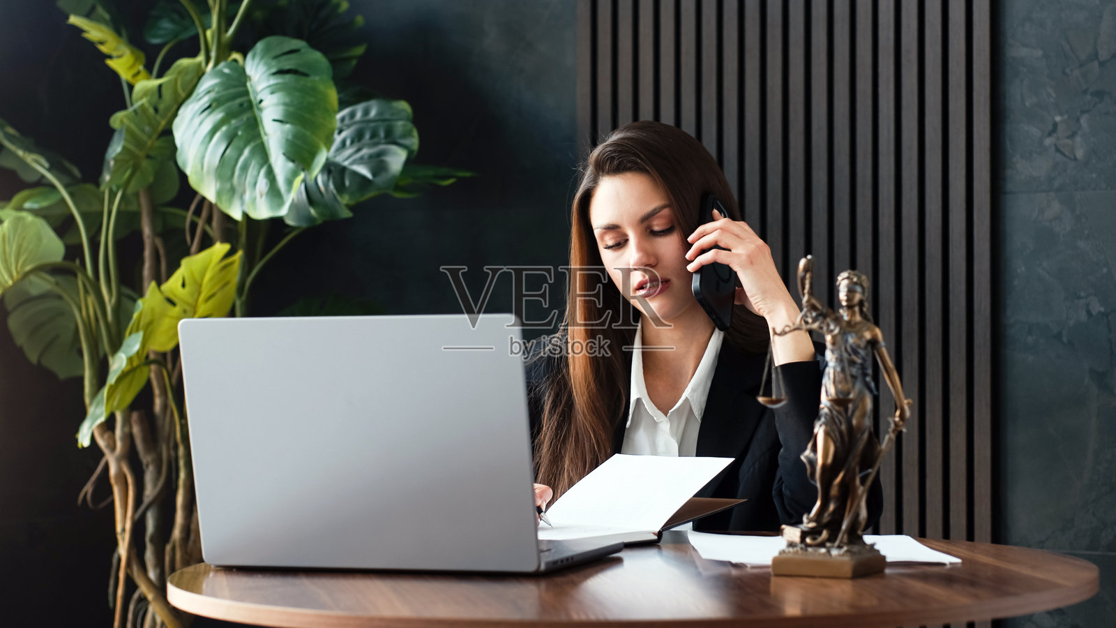 女性律师专注于在木桌上写笔记，桌上有一台笔记本电脑和一尊女神法像。照片摄影图片