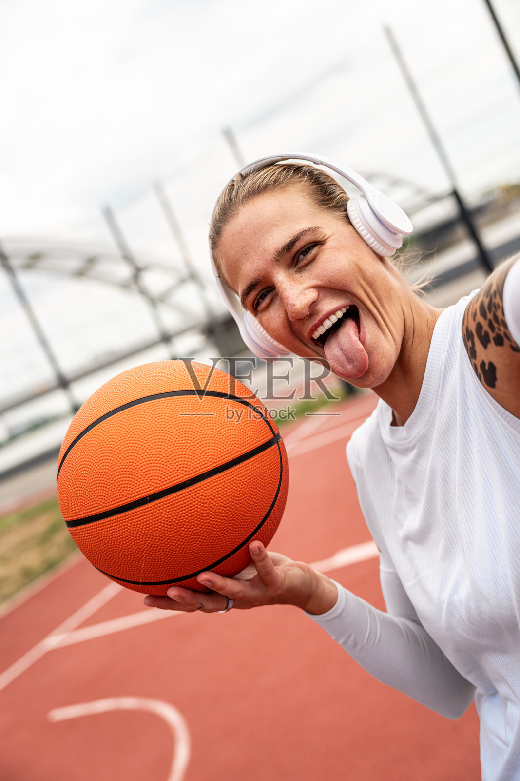年轻女性佩戴耳机，手持篮球，展现酷炫态度的肖像，背景为城市运动场。照片摄影图片