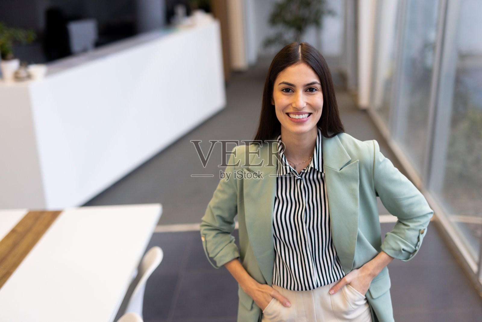 微笑的女商人在现代办公室自信地站立，双手插在口袋里，散发着积极和专业的优雅气质照片摄影图片