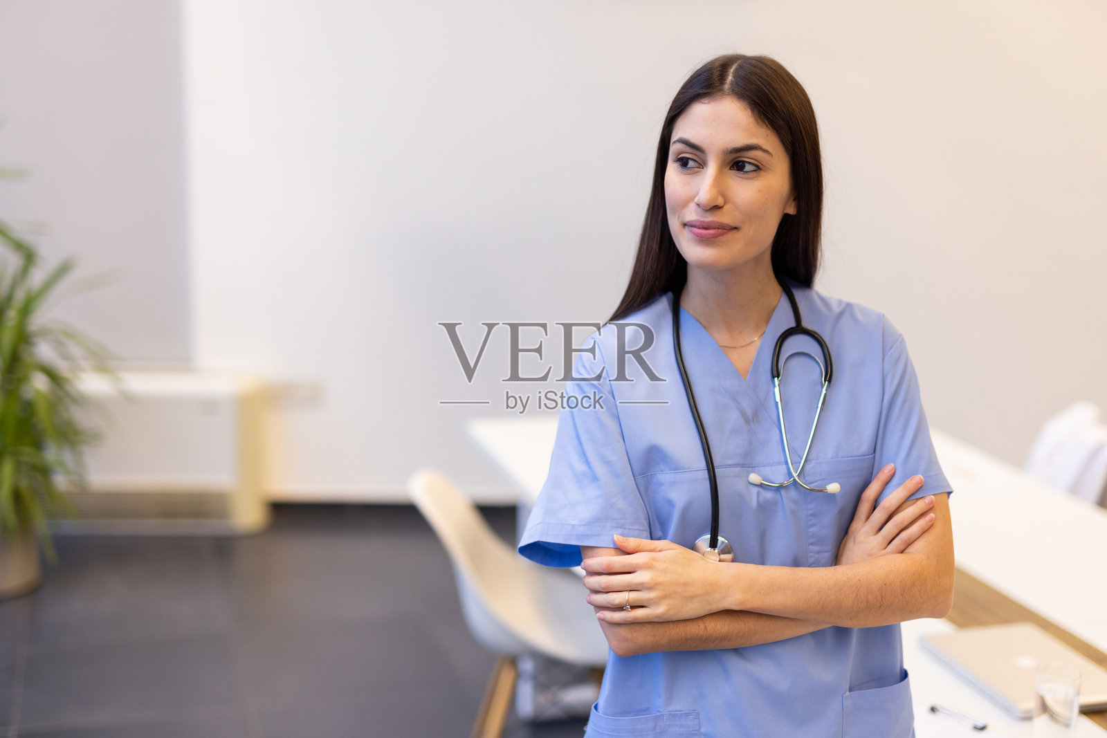 穿着蓝色医务服的女性医生，自信地交叉双臂，站在现代医院环境中的医疗设备旁。照片摄影图片