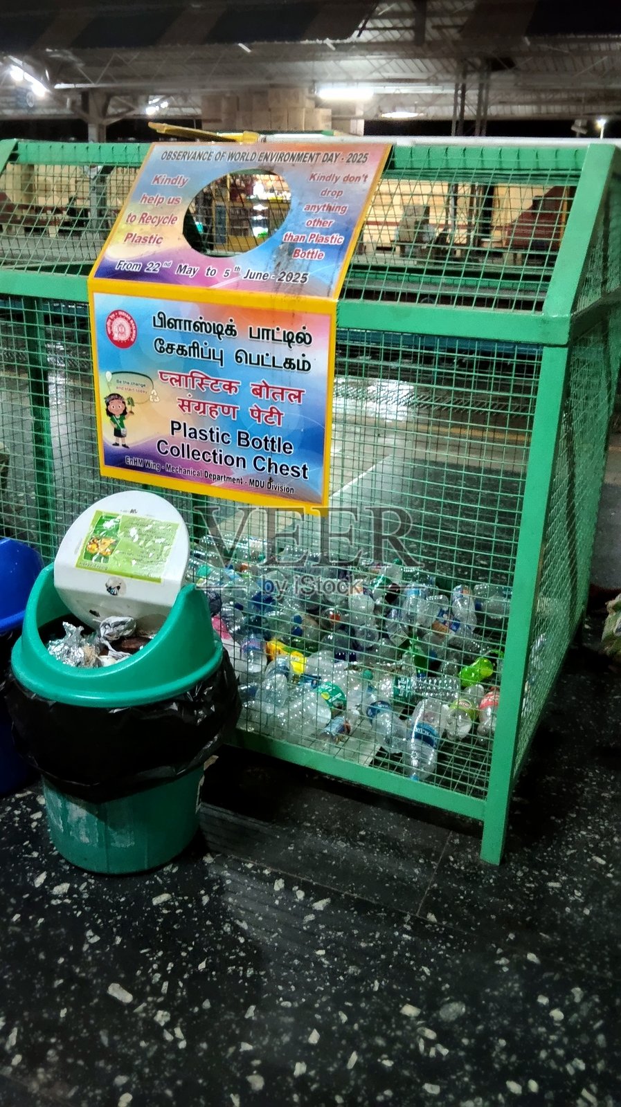 一个用于收集塑料瓶的金属笼，一个绿色垃圾桶，以及一个宣传塑料回收的横幅，位于马杜赖南方铁路车站，庆祝世界环境日。照片摄影图片