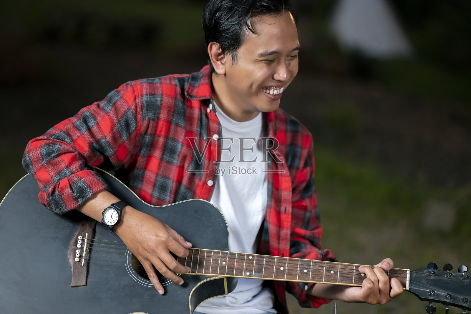 东南亚印尼男子在户外弹奏原声吉他，展现轻松愉悦的生活方式。享受自然环境中露营生活的场景。照片摄影图片