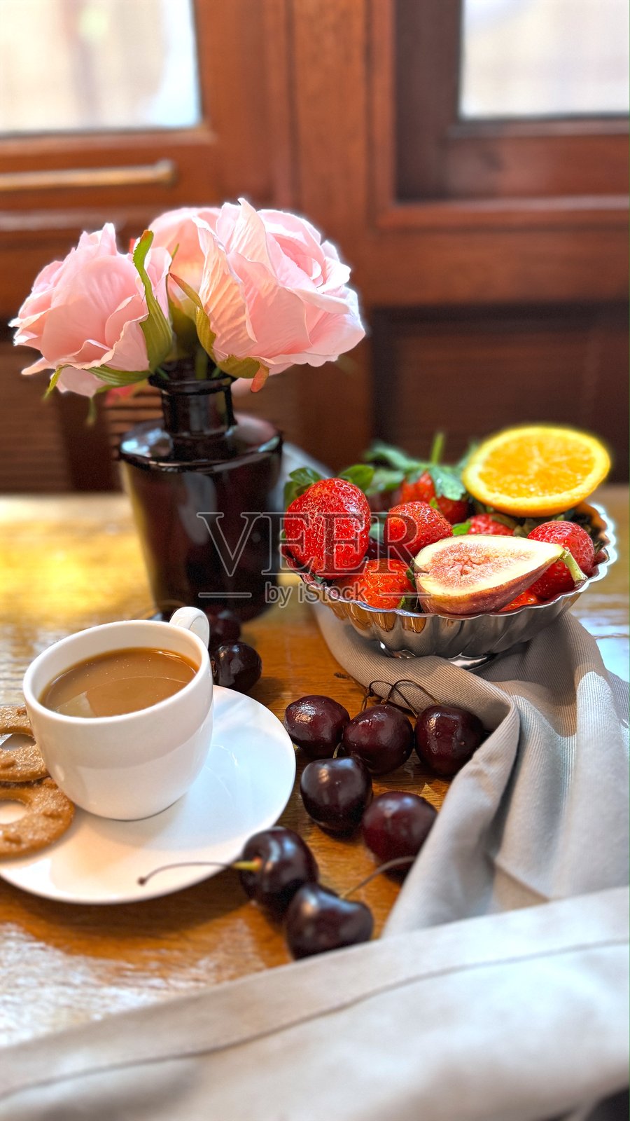 白色咖啡杯里装满牛奶，旁边放着饼干，木桌上还有一碗新鲜的草莓、无花果和橙子片，旁边点缀着粉色玫瑰。照片摄影图片